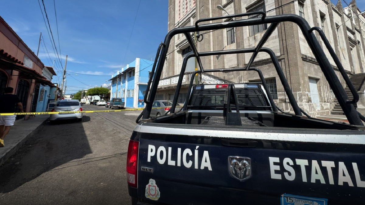 Vista sobre la batea de una camioneta de POLICÍA ESTATAL en la calle de una ciudad colonial (Colima). La calle está acordonada con cinta amarilla, y se ven edif