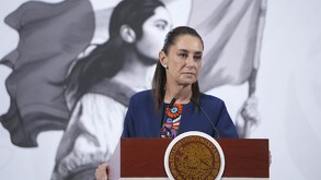 La política Claudia Sheinbaum (con un traje azul marino y una blusa bordada de colores) está detrás de un podio con el escudo de México