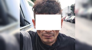 Hombre detenido viendo de frente con los ojos censurados, vistiendo una playera negra con una calle de fondo y automóviles estacionados a los costados