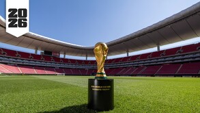 El Trofeo de la Copa Mundial de la FIFA sobre su base en el césped del Estadio Akron (Guadalajara). Las gradas rojas vacías rodean el campo. El logo de 