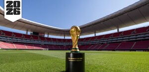 El Trofeo de la Copa Mundial de la FIFA sobre su base en el césped del Estadio Akron (Guadalajara). Las gradas rojas vacías rodean el campo. El logo de 
