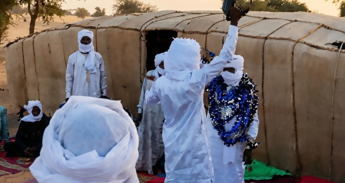 Hombre vestido con tunica blanca levantando el brazo mientras sostiene una ametralladora con personas vestidas igual que él alrededor