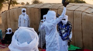 Hombre vestido con tunica blanca levantando el brazo mientras sostiene una ametralladora con personas vestidas igual que él alrededor