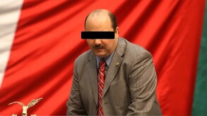 César Duarte, ex gobernador de Chihuahua, mirando a la cámara. Viste un traje gris y una camisa blanca. De fondo el color rojo en la bandera.