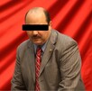 César Duarte, ex gobernador de Chihuahua, mirando a la cámara. Viste un traje gris y una camisa blanca. De fondo el color rojo en la bandera.