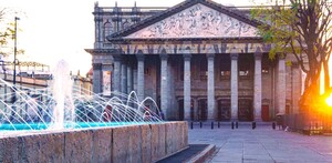 El histórico Teatro Degollado en Guadalajara. La fachada neoclásica tiene columnas y un relieve. Una fuente moderna con chorros iluminados en azul