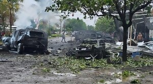 Múltiples automóviles calcinados con ramas esparcidas alrededor y un par de personas caminando en la zona donde un coche bomba explotó