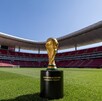 El Trofeo de la Copa Mundial de la FIFA sobre su base en el césped del Estadio Akron (Guadalajara). Las gradas rojas vacías rodean el campo. El logo de 