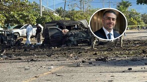 Una escena de destrucción y escombros en una carretera, con los restos quemados de varios vehículos. Hay personal de investigación en trajes protectores blancos