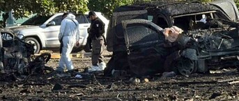 Una escena de destrucción y escombros en una carretera, con los restos quemados de varios vehículos. Hay personal de investigación en trajes protectores blancos