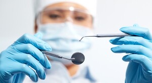 Mujer dentista usando gafas, cubrebocas y guantes mientras sostiene herramientas odontológicas en ambas manos