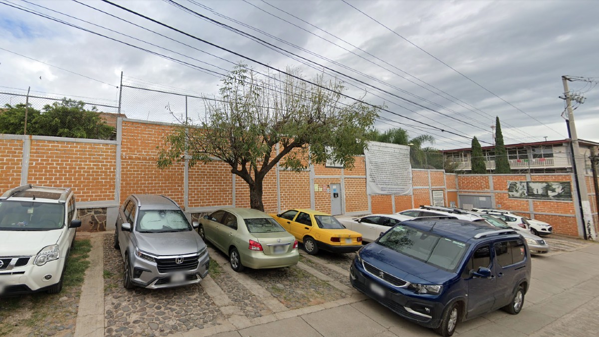 Un lote de estacionamiento exterior con varios autos (incluyendo un Nissan, Honda, Toyota y un auto amarillo tipo taxi) estacionados frente a una larga barda de