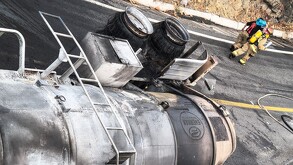 imagen dramática de un camión cisterna que ha volcado y está ardiendo en una curva de una carretera montañosa. Muestra la respuesta de emergencia con humo