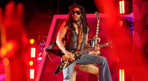 Lenny Kravitz sobre un escenario tocando una guitarra eléctrica frente a una multitud con las manos arriba ambientado con luces rojas