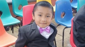 Niño vestido de traje y moño, sentado sobre una silla en color naranja.