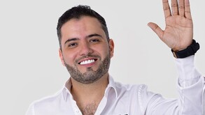 Primer plano de un hombre sonriente con barba, cabello oscuro y una camisa blanca abotonada. Levanta la mano derecha en un gesto de saludo o despedida.