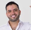 Primer plano de un hombre sonriente con barba, cabello oscuro y una camisa blanca abotonada. Levanta la mano derecha en un gesto de saludo o despedida.