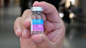 Primer plano de una mano sosteniendo un pequeño vial de vidrio con una tapa plateada que contiene la Vacuna Antigripal 