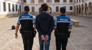 Joven vistiendo sudadera azul y con las manos esposadas siendo arrestado por dos agentes de policía en el medio de un patio en España