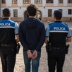 Joven vistiendo sudadera azul y con las manos esposadas siendo arrestado por dos agentes de policía en el medio de un patio en España