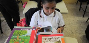 Se observa a una niña con un libro dentro de un salón de clases.