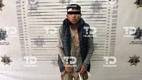 Un hombre con ojos tapados que usa una blusa gris y un chaleco negro.