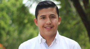 Enrique Estrada Jiménez viendo de frente, sonriendo y vistiendo una camisa blanca con un fondo desenfocado de plantas verdes