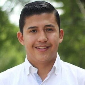 Enrique Estrada Jiménez viendo de frente, sonriendo y vistiendo una camisa blanca con un fondo desenfocado de plantas verdes