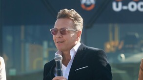 Pablo Lemus (saco oscuro, camisa blanca y gafas de sol rosas) habla en un micrófono en un evento exterior. Se distingue un fondo azul oscuro