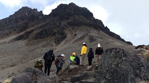 Un grupo de alpinistas (siete personas) se reúne al pie de una montaña rocosa oscura y empinada, bajo un cielo nublado. Están de pie sobre rocas más pequeñas