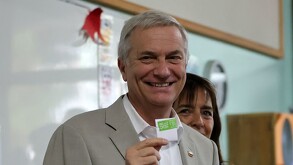 Primer plano del expresidente chilenoJosé Antonio Kast (pelo gris, traje claro, camisa blanca) sonriendo, mientras sostiene una papeleta de votación. Una mujer