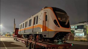 Un vagón moderno de tren, de color blanco y anaranjado, es transportado sobre un tráiler de plataforma por la noche. Este tren es parte de la flota
