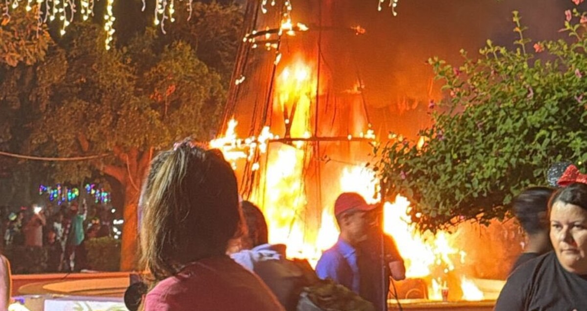 Estructura de pino de Navidad ardiendo en llamas con personas caminando y observando con árboles y luces navideñas alrededor