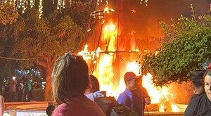 Estructura de pino de Navidad ardiendo en llamas con personas caminando y observando con árboles y luces navideñas alrededor