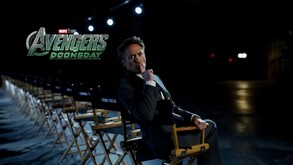 El actor Robert Downey Jr. (traje oscuro, mano en la barbilla) está sentado en una silla de director en un estudio oscuro. En la esquina superior izquierda se l