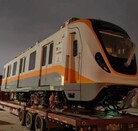 Un vagón moderno de tren, de color blanco y anaranjado, es transportado sobre un tráiler de plataforma por la noche. Este tren es parte de la flota