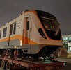 Un vagón moderno de tren, de color blanco y anaranjado, es transportado sobre un tráiler de plataforma por la noche. Este tren es parte de la flota
