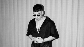 Malilla posa en blanco y negro, con gafas de sol oscuras y una camisa negra de manga corta, abierta. Lleva un gran collar de cruz y un reloj.