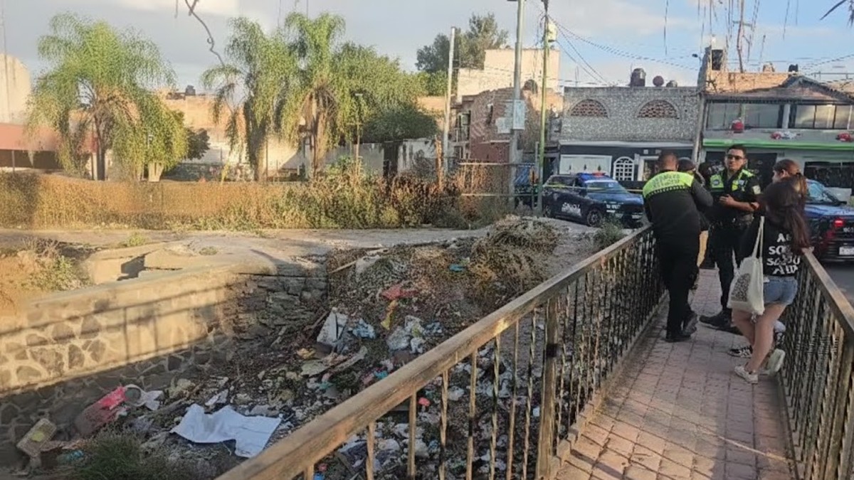Dos agentes de policía se encuentran en un puente sobre un canal abierto lleno de basura y vegetación. Una mujer con una camiseta blanca está a su lado.
