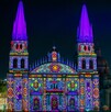 La Catedral de Guadalajara con sus torres gemelas está iluminada de noche con un patrón de luces de colores brillantes como magenta, verde, amarillo y azul.