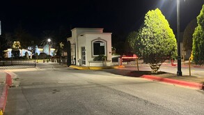 Se observan un camino, unos árboles, un portón negro y una caseta de vigilancia.