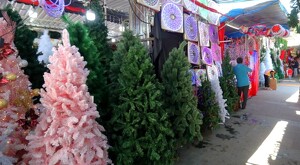 Árboles de Navidad de distintos colores y tamaños exhibidos junto a luces y adornos navideños en medio de un tianguis con personas y vendedores alrededor