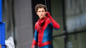 El actor Tom Holland posa como Spider-Man con el traje rojo y azul, pero sin la máscara puesta. Sostiene un hilo de telaraña con su mano derecha.