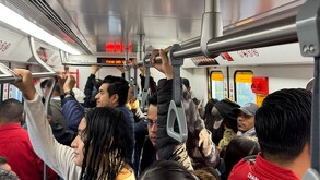 Un tren moderno blanco y naranja de la Línea 4 está en la estación. Una oficial vigila en el andén mientras pasajeros se ven dentro del vagón.