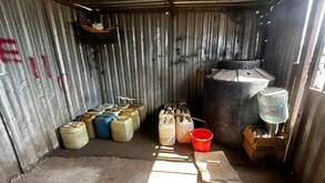 Interior de una bodega de lámina con numerosos bidones de plástico, un tinaco negro grande, una cubeta roja y un garrafón de agua sobre cajas.