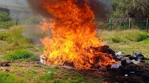 Una gran hoguera con llamas intensas de color naranja y humo negro en un campo abierto. Al lado del fuego se ven cajas y bultos siendo incinerados.