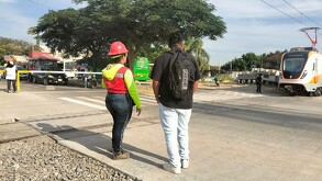 Una trabajadora con casco rojo y un joven con mochila observan el paso de un tren moderno de la Línea 4 en un cruce vial con autos y árboles.