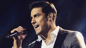 Se observa a un hombre con micrófono en mano cantando, viste un saco gris.