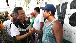 Pablo Lemus Navarro estrecha la mano de un hombre con camiseta de tirantes frente a un muro blanco. Hay personas y militares en el fondo.