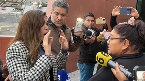 Se observa a una mujer hablando, viste saco blanco con negro y alrededor de ella hay varios hombres con micrófonos.